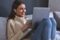 Улыбающаяся белая женщина проводит время дома, сидя на диване в гостиной, используя ноутбук с наушниками, держа кружку. Социальное дистанцирование во время изоляции коронавируса Covid 19. — стоковое фото