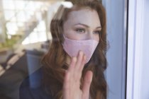 Uma mulher caucasiana atraente, gengibre, passando tempo em casa, na sala de estar, olhando pela janela, usando uma máscara facial. Distanciamento social durante o bloqueio de quarentena do Covid 19 Coronavirus. — Fotografia de Stock