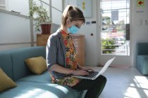 Белая деловая женщина, сидящая на диване в офисе в маске для лица с помощью ноутбука. Здоровье и гиперактивность на рабочем месте во время коронавируса Ковид 19 пандемии. — стоковое фото