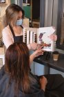 Белая женщина-парикмахер работает в парикмахерской в маске для лица, показывая красители для волос белой женщине. Здоровье и гиперактивность на рабочем месте во время коронавируса Ковид 19 пандемии. — стоковое фото