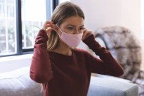 Mujer caucásica pasando tiempo en casa, sentada en la sala de estar, poniéndose mascarilla. Distanciamiento social durante el bloqueo de cuarentena del Coronavirus Covid 19 - foto de stock