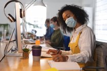 Frauen mit gemischter Rasse sitzen kreativ am Schreibtisch in einem Büro mit Gesichtsmaske, benutzen einen Computer und schreiben in ein Notizbuch. Gesundheit und Hygiene am Arbeitsplatz während der Coronavirus Covid 19 Pandemie. — Stockfoto
