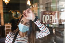 Kaukasische Friseurin, die im Friseursalon arbeitet, trägt Gesichtsmaske und lehnt an einer Glastür mit einem Schild, auf dem steht, dass sie offen ist. Gesundheit und Hygiene am Arbeitsplatz während der Coronavirus Covid 19 Pandemie. — Stockfoto