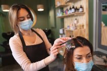 Белая женщина-парикмахер работает в парикмахерской в маске для лица, вымирающие волосы белой клиентки в маске. Здоровье и гиперактивность на рабочем месте во время коронавируса Ковид 19 пандемии. — стоковое фото
