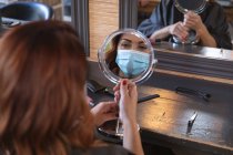 Femme caucasienne cliente d'un coiffeur portant un masque facial et regardant son reflet dans le miroir. Santé et hygiène sur le lieu de travail pendant la pandémie de Coronavirus Covid 19. — Photo de stock