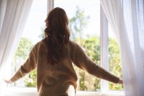 Вид сзади на рыжую белую женщину, проводящую время дома, в гостиной, смотрящую в окно. Социальное дистанцирование во время изоляции коронавируса Covid 19. — стоковое фото