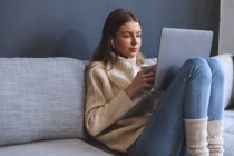 Белая женщина проводит время дома, сидя на диване в гостиной, используя ноутбук с наушниками, держа кружку. Социальное дистанцирование во время изоляции коронавируса Covid 19. — стоковое фото