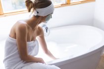 Caucasian woman spending time at home, in bathroom with face mask on, running bath sitting on edge of bathtub. Social distancing during Covid 19 Coronavirus quarantine lockdown. — Stock Photo