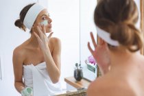 Кавказька жінка проводить час удома, стоїть у ванній кімнаті і дивиться у дзеркало, застосовуючи маску обличчя. Соціальна дистанція в Ковиді 19 Коронавірус карантин. — стокове фото