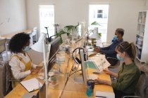Grupo multiétnico de creativos masculinos y femeninos que trabajan en escritorios de oficina con pantallas protectoras, utilizando computadoras. Salud e higiene en el lugar de trabajo durante la pandemia de Coronavirus Covid 19. - foto de stock