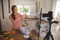 Caucasian woman spending time at home, putting fruit in a blender, recording it with a camera. Social distancing during Covid 19 Coronavirus quarantine lockdown. — Stock Photo