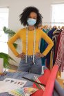 Porträt einer Modedesignerin mit gemischter Rasse im Studio, die Gesichtsmaske und Maßband um den Hals trägt und in die Kamera blickt. Gesundheit und Hygiene am Arbeitsplatz während der Coronavirus Covid 19 Pandemie. — Stockfoto