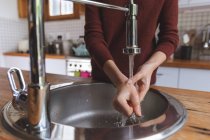 Sección media de la mujer que pasa tiempo en casa, de pie en la cocina lavándose las manos en el lavabo. Distanciamiento social durante el bloqueo de cuarentena del Coronavirus Covid 19. - foto de stock
