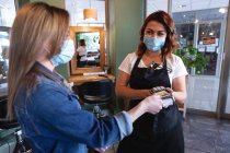 Белая женщина-парикмахер, работающая в парикмахерской в маске для лица, получает оплату от клиентки в маске. Здоровье и гиперактивность на рабочем месте во время коронавируса Ковид 19 пандемии. — стоковое фото