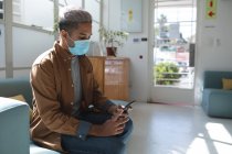Mixed race male business creative sitting on sofa in an office wearing face mask using his smartphone. Health and hygiene in workplace during Coronavirus Covid 19 pandemic. — Stock Photo