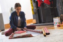Caucasian woman spending time at home at Christmas, sitting on floor by fireplace in living room, cutting wrapping paper. Social distancing during Covid 19 Coronavirus quarantine lockdown. — Stock Photo