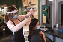 Белая женщина-парикмахер работает в парикмахерской в маске для лица, расчесывая волосы белой клиентки в маске для лица. Здоровье и гиперактивность на рабочем месте во время коронавируса Ковид 19 пандемии. — стоковое фото