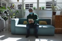 Homme d'affaires mixte créatif assis sur un canapé dans un bureau portant un masque facial à l'aide de sa tablette. Santé et hygiène sur le lieu de travail pendant la pandémie de Coronavirus Covid 19. — Photo de stock