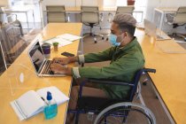 Gemischte Rassenmännchen, die im Rollstuhl am Schreibtisch in einem Büro sitzen, Gesichtsmaske tragen und einen Laptop benutzen. Gesundheit und Hygiene am Arbeitsplatz während der Coronavirus Covid 19 Pandemie. — Stockfoto