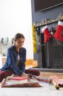 Femme caucasienne passant du temps à la maison à Noël, assise sur le sol près de la cheminée dans le salon, coupant du papier d'emballage. Distance sociale pendant le confinement en quarantaine du coronavirus Covid 19. — Photo de stock