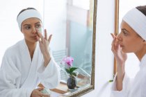Donna caucasica trascorrere del tempo a casa, in piedi in bagno, guardando nello specchio applicare maschera viso. Distanza sociale durante il blocco di quarantena Covid 19 Coronavirus. — Foto stock