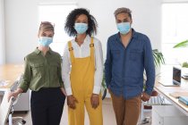 Retrato de un grupo multiétnico de tres creativos masculinos y femeninos en el cargo con máscaras faciales, Salud e higiene en el lugar de trabajo durante la pandemia de Coronavirus Covid 19. - foto de stock