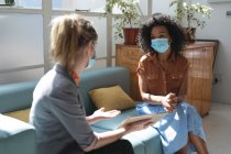 Mixed race and Caucasian female business creatives wearing face masks and distancing on sofa, talking and using tablet in office. Health and hygiene in workplace during Coronavirus Covid 19 pandemic. — Stock Photo