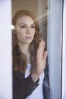 Une jolie femme caucasienne rousse passe du temps à la maison, dans le salon, regardant par la fenêtre. Distance sociale pendant le confinement en quarantaine du coronavirus Covid 19. — Photo de stock