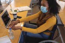 Mixed race female creative sitting in a wheelchair at desk in an office, wearing face mask, disinfecting hands with hand santiser. Health and hygiene in workplace during Coronavirus Covid 19 pandemic. — Stock Photo