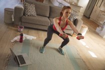 Caucasian woman spending time at home, in living room, exercising with dumbbells, using laptop. Social distancing during Covid 19 Coronavirus quarantine lockdown. — Stock Photo