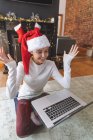 Mujer caucásica feliz pasar tiempo en casa en Navidad, con sombrero de Santa, sentado junto a la mesa utilizando el ordenador durante el chat de vídeo. Distanciamiento social durante el bloqueo de cuarentena del Coronavirus Covid 19. - foto de stock