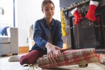 Mujer caucásica pasar tiempo en casa en Navidad, sentado en el suelo junto a la chimenea en la sala de estar, envolviendo presente en papel. Distanciamiento social durante el bloqueo de cuarentena del Coronavirus Covid 19. - foto de stock