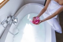 Mid section of woman spending time at home, in bathroom holding rose petals, sitting on edge of bathtub. Social distancing during Covid 19 Coronavirus quarantine lockdown. — Stock Photo