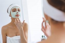 Donna caucasica trascorrere del tempo a casa, in piedi in bagno, guardando nello specchio applicare maschera viso. Distanza sociale durante il blocco di quarantena Covid 19 Coronavirus. — Foto stock