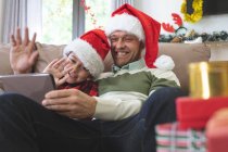 Hombre caucásico en casa con su hijo en Navidad, con sombreros de Santa sentado en el sofá en la sala de estar, usando tableta digital, saludando. Distanciamiento social durante el bloqueo de cuarentena del Coronavirus Covid 19. - foto de stock