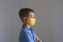 Vue latérale du garçon caucasien passant du temps à la maison, portant un masque jaune regardant la caméra sur fond gris. Distance sociale pendant le confinement en quarantaine du coronavirus Covid 19. — Photo de stock
