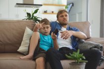 Kaukasischer Mann zu Hause mit seinem Sohn zusammen, auf dem Sofa im Wohnzimmer sitzend, vor dem Fernseher. Soziale Distanzierung während Covid 19 Coronavirus Quarantäne Lockdown. — Stockfoto