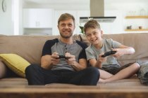 Homme caucasien à la maison avec son fils ensemble, assis sur le canapé dans le salon, jouant à des jeux vidéo, souriant. Distance sociale pendant le confinement en quarantaine du coronavirus Covid 19. — Photo de stock