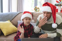 Hombre caucásico en casa con su hijo en Navidad, con sombreros de Santa sentado en la sala de estar utilizando el ordenador portátil para videollamada. Distanciamiento social durante el bloqueo de cuarentena del Coronavirus Covid 19. - foto de stock