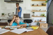 Белый мужчина дома со своим сыном вместе, на кухне, мальчик делает домашнее задание за столом, думает. Социальное дистанцирование во время изоляции коронавируса Covid 19. — стоковое фото