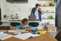 Белый мужчина дома со своим сыном вместе, на кухне, мальчик делает домашнее задание за столом. Социальное дистанцирование во время изоляции коронавируса Covid 19. — стоковое фото