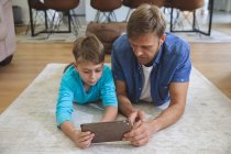 Homem caucasiano em casa com seu filho junto, deitado no tapete na sala de estar, usando tablet digital. Distanciamento social durante o bloqueio de quarentena do Covid 19 Coronavirus. — Fotografia de Stock