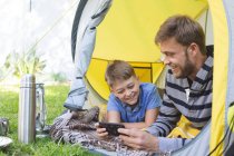 Caucasian man spending time with his son together, camping in garden, lying in tent using smartphone, smiling. Social distancing during Covid 19 Coronavirus quarantine lockdown. — Stock Photo