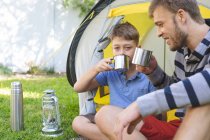 Caucasian man spending time with his son together, camping in garden, sitting next to tent drinking tea, smiling. Social distancing during Covid 19 Coronavirus quarantine lockdown. — Stock Photo