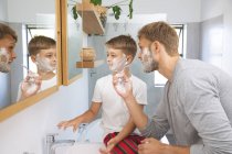 Homme caucasien à la maison avec son fils ensemble, dans la salle de bain, rasage avec de la crème à raser sur les visages, regarder le miroir. Distance sociale pendant le confinement en quarantaine du coronavirus Covid 19. — Photo de stock