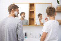 Hombre caucásico en casa con su hijo juntos, en el baño, afeitándose con crema de afeitar en las caras, mirando al espejo. Distanciamiento social durante el bloqueo de cuarentena del Coronavirus Covid 19. - foto de stock