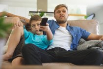 Hombre caucásico en casa con su hijo juntos, sentado en el sofá en la sala de estar, viendo la televisión. Distanciamiento social durante el bloqueo de cuarentena del Coronavirus Covid 19. - foto de stock