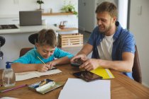 Caucasian man at home with his son together, in kitchen, father helping the boy doing homework at table. Social distancing during Covid 19 Coronavirus quarantine lockdown. — Stock Photo