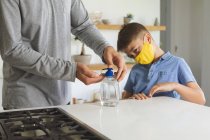 Caucasian man at home with his son, in kitchen, boy wearing face mask, man sanitizing his hands. Social distancing during Covid 19 Coronavirus quarantine lockdown. — Stock Photo