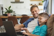 Hombre caucásico en casa con su hijo juntos, sentado en el sofá en la sala de estar, utilizando el ordenador portátil, sonriendo. Distanciamiento social durante el bloqueo de cuarentena del Coronavirus Covid 19. - foto de stock